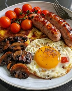 a white plate topped with sausages, eggs and mushrooms next to tomatoes on a table