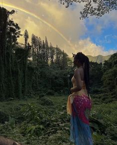 a woman standing in the middle of a forest with a rainbow in the sky behind her