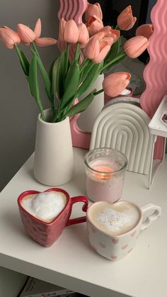 pink tulips in a vase and two mugs on a white table next to each other