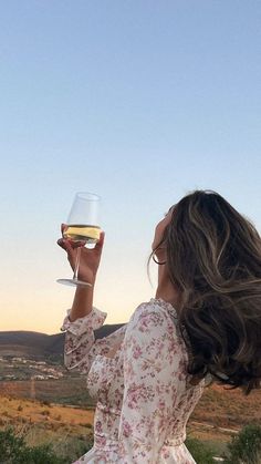 a woman holding a wine glass up to her face while sitting on a hill top