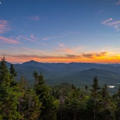 the sun is setting over mountains and trees in the foreground, as seen from an overlook