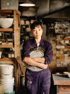 a woman standing in a room with lots of pots on the wall and she has her arms crossed