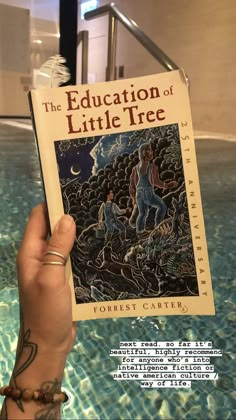 a person holding up a book in front of a swimming pool with the words education of little tree written on it