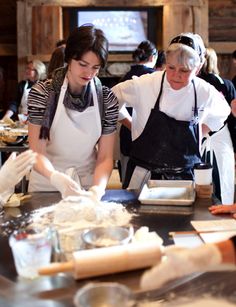 two women in aprons are preparing food on a table with other people behind them