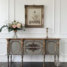 an antique sideboard with two drawers and flowers in vase next to the painting on the wall