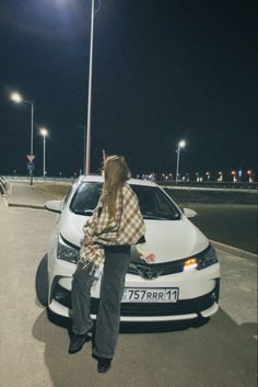 a woman sitting on the hood of a white car in a parking lot at night