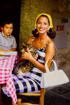 a woman holding a cat while sitting at a table with a boy in the background