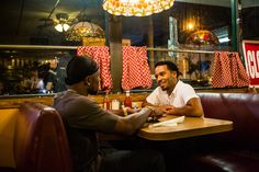 two men sitting at a table talking to each other in a restaurant with red and white banners hanging on the wall