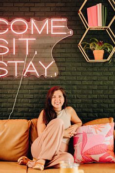 a woman sitting on a couch in front of a neon sign that says come sit stay