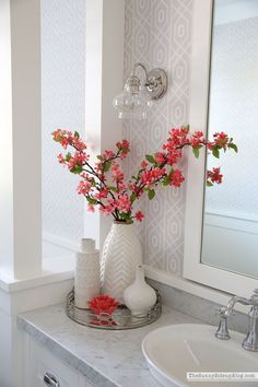 a white vase filled with pink flowers sitting on top of a bathroom counter next to a mirror