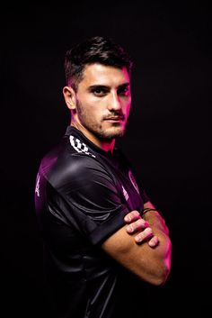 a man with his arms crossed standing in front of a black background wearing a purple shirt