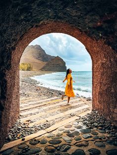 a woman in a yellow dress is walking through an archway to the ocean and mountains