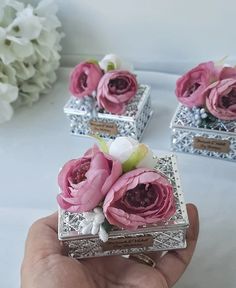 a hand is holding three small silver boxes with pink flowers in them on a table