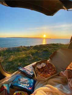 the sun is setting over an open car trunk with pizza and chips on it, along with snacks