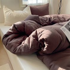 an unmade bed with a book on top of it next to pillows and a window