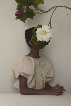 a woman with flowers on her head sitting in front of a white wall next to a plant