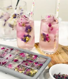 two glasses filled with ice and flowers on top of a wooden cutting board next to some drinks