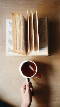 a person holding a cup with tea in it and an open book on the table