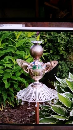 a silver vase sitting on top of a wooden stand in front of green plants and bushes