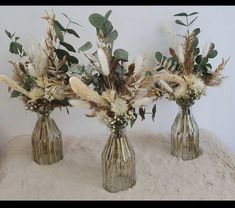 three vases with dried flowers and greenery in them on a white blanketed surface