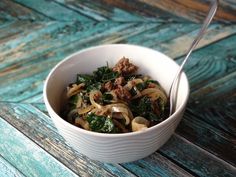 a white bowl filled with pasta and meat on top of a wooden table next to a spoon