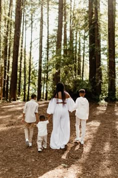 a woman and two children walking through the woods in white outfits with trees behind them