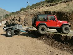 a red jeep pulling a trailer down a dirt road on the side of a hill