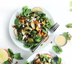 two plates filled with salad and dressing on top of a white table next to silverware