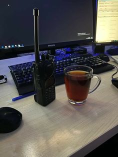 a cup of tea next to a cell phone on a desk with a computer monitor and keyboard