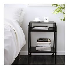 a black end table with some books on it next to a white bed and plant