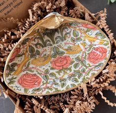 a decorative bowl sitting on top of wood shavings in a box with flowers and birds painted on it