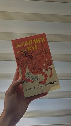 the catcher in the rye book being held by a person's hand with blinds behind it