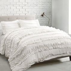 a white bed with ruffles and pillows in a room next to a brick wall