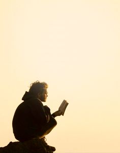 a person sitting on top of a rock reading a book while the sun is setting