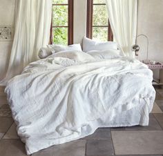 an unmade bed with white linens and pillows in front of two large windows