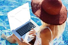 a woman sitting on the edge of a swimming pool using a laptop computer while wearing a straw hat