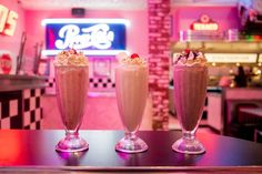 three drinks sitting on top of a table in front of a neon lit restaurant sign