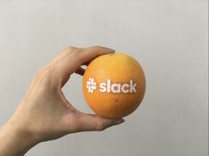 a hand holding an orange with the word slack on it in front of a white wall