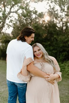 a pregnant woman hugging her husband while standing in front of some trees and grass at sunset