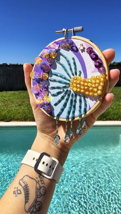 a person holding up a small embroidery piece in front of a swimming pool with grass and blue sky