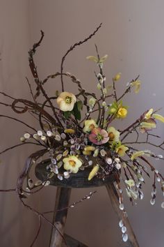 an arrangement of flowers and branches in a vase on a wooden stand against a wall