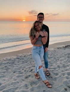 a man and woman are standing on the beach at sunset with their arms around each other