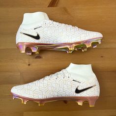 a pair of white and pink nike soccer cleats on a wooden floor with gold accents