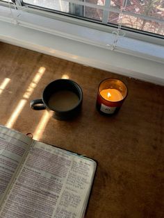 a cup of coffee next to an open book and candle on a table in front of a window