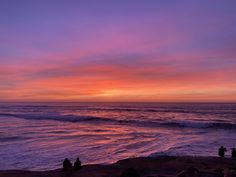 the sun is setting over the ocean and people are sitting on the rocks looking out at the water