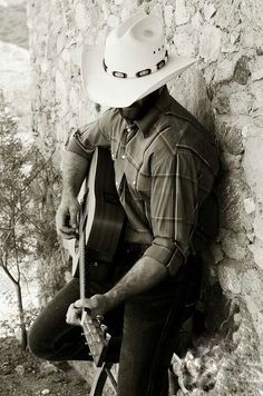 a man wearing a cowboy hat and holding a guitar leaning against a stone wall with trees in the background