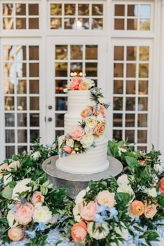 a white wedding cake surrounded by flowers and greenery on a table in front of doors