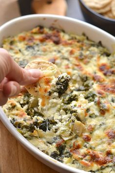 a hand holding a tortilla chip over a casserole filled with spinach and cheese