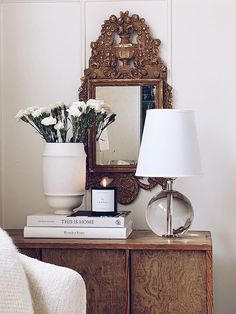 a table with flowers and a lamp on it in front of a mirror, next to a chair