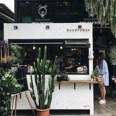 a woman standing in front of a white building with plants on the outside and people behind it